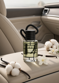 A clear glass perfume bottle labeled 'RP LUXE DREAM' with a black cap and black cord, placed on a beige car seat with cotton bolls, vanilla beans, and white flowers around it.