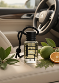 A clear glass car fragrance bottle labeled RP BALE QALDER with a black cap and hanging cord, styled on a beige car seat with green leaves, a lime, and an orange slice nearby. The blurred car interior and steering wheel are in the background.