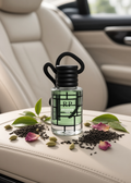 A clear glass car fragrance bottle labeled 'RP London Tea' with green tea leaves inside, placed on a beige leather car seat. The bottle is styled with scattered tea leaves, green cardamom pods, and pink rose petals.
