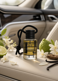 A clear glass perfume bottle with a black cap and black hanging cord sits on a beige leather car seat, surrounded by white flowers, a lime half, a green bumpy fruit, and dark vanilla beans. The bottle label reads RP and CASALANICA.