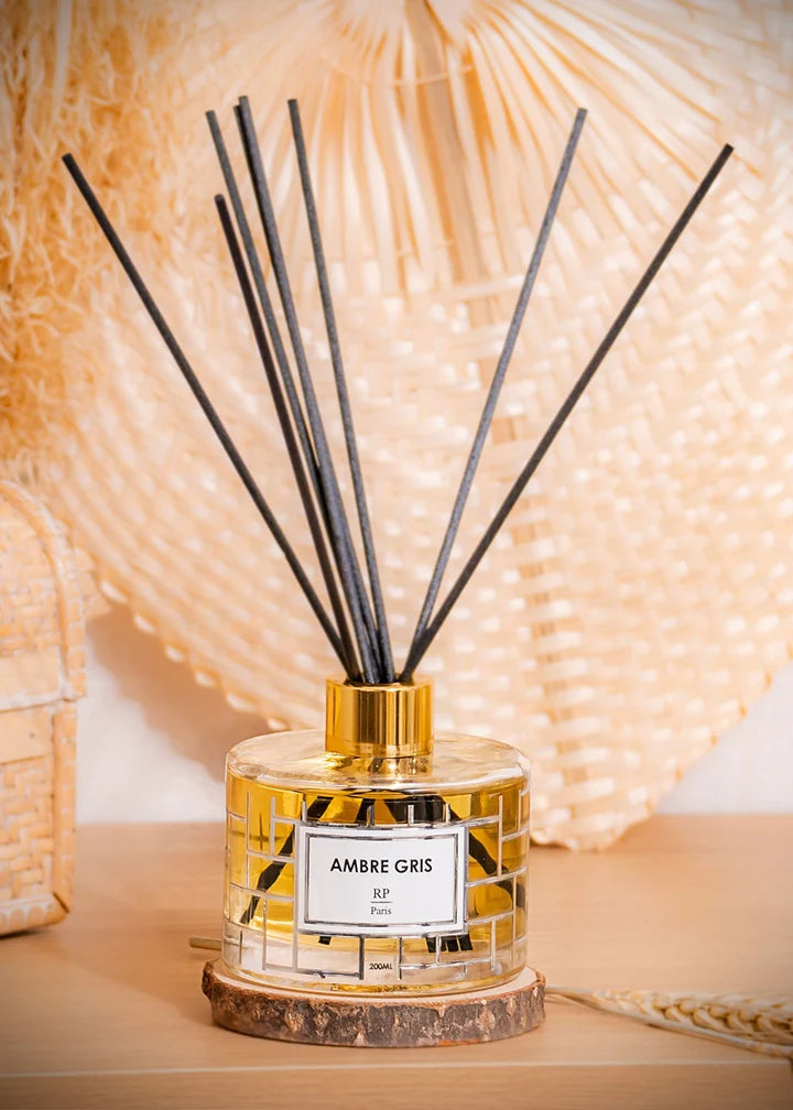 A reed diffuser bottle with a gold cap and black reeds, labeled 'AMBRE GRIS', set on a wooden slice coaster against a warm beige woven background.