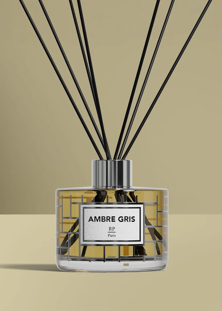 A reed diffuser bottle labeled 'AMBRE GRIS' with a white label and black reeds, set against a warm beige background.