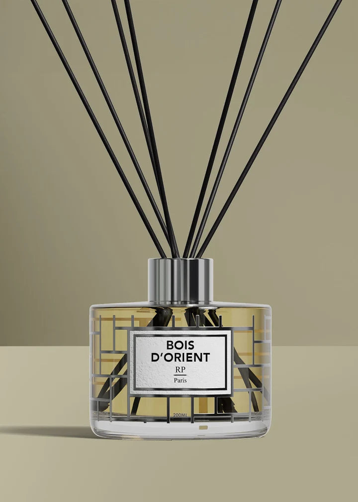 A reed diffuser bottle with black reeds, a clear glass body, and a white label reading 'BOIS D'ORIENT JP Paris', set against a warm beige background.