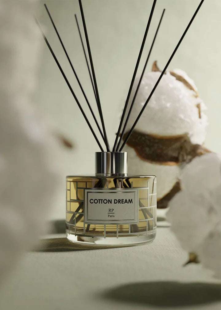 A reed diffuser bottle labeled 'COTTON DREAM' with several black reed sticks, styled with cotton bolls on a soft neutral background.