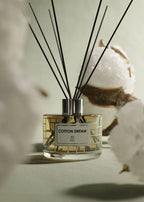 A reed diffuser bottle labeled 'COTTON DREAM' with several black reed sticks, styled with cotton bolls on a soft neutral background.
