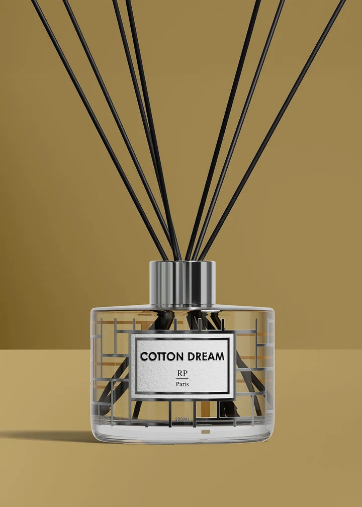 A reed diffuser bottle with black reeds, a clear glass body, and a white label reading 'COTTON DREAM' and 'RP Paris', set against a warm tan background.