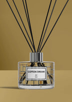 A reed diffuser bottle with black reeds, a clear glass body, and a white label reading 'COTTON DREAM' and 'RP Paris', set against a warm tan background.