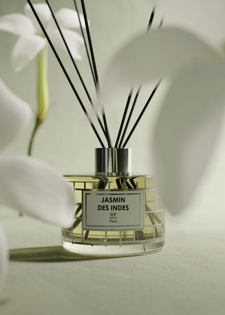 A reed diffuser bottle with black reeds and a clear faceted glass base, labeled 'JASMIN DES INDES EP Paris,' styled with soft white flowers in the foreground and background.