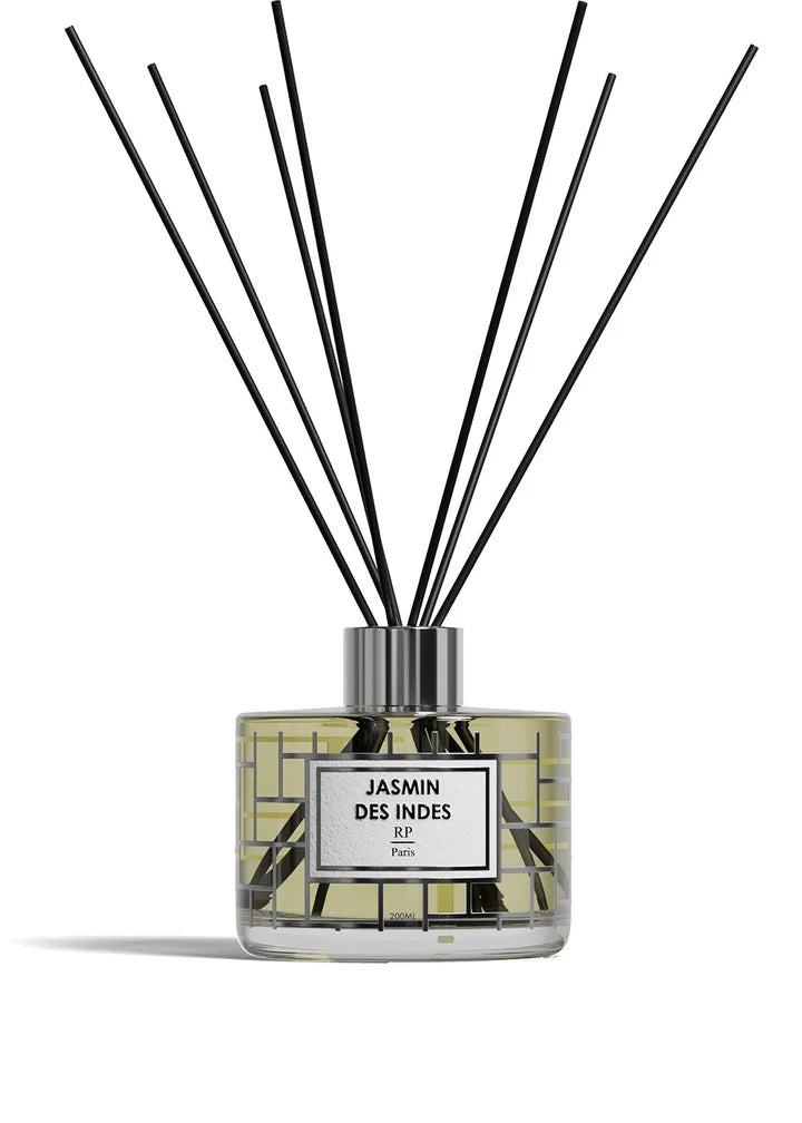 A reed diffuser bottle with a clear glass body, pale yellow liquid, a white label reading 'ROSE DE MAI BP Paris,' and multiple black reed sticks extending upward against a white background.