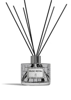 A reed diffuser bottle with a clear glass body, pale yellow liquid, a white label reading 'ROSE DE MAI BP Paris,' and multiple black reed sticks extending upward against a white background.