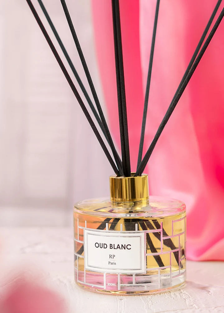 A reed diffuser bottle with black reeds, a gold cap, and a white label reading 'OUD BLANC RP Paris', set against a soft pink and white background.
