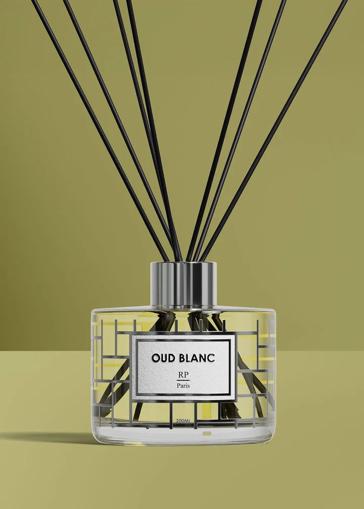 A clear glass reed diffuser bottle with a white label reading 'OUD BLANC' and 'RP Paris', filled with pale yellow liquid, topped with a silver cap and black reeds extending upward against a muted olive-green background.