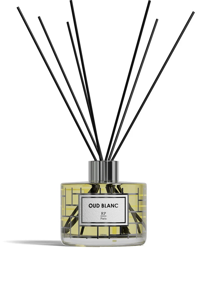 A reed diffuser bottle with a clear glass body, pale yellow liquid, a white label reading 'ROSE DE MAI BP Paris,' and multiple black reed sticks extending upward against a white background.