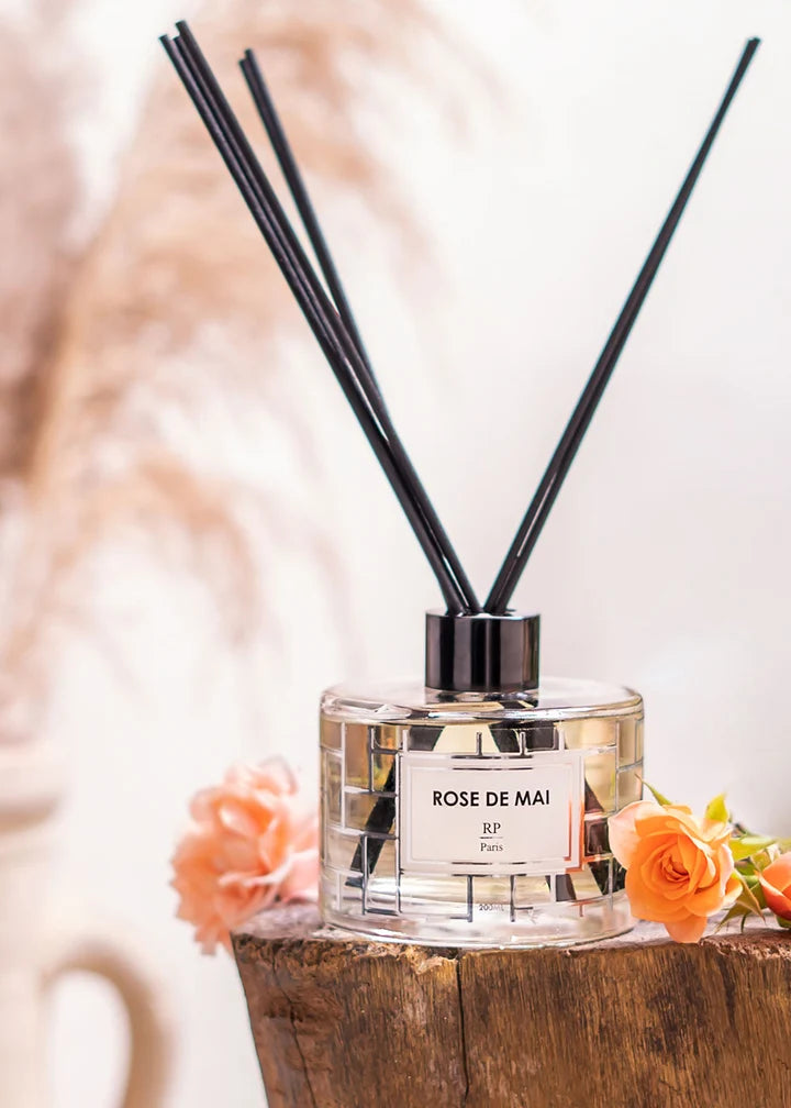 A clear glass reed diffuser bottle labeled 'ROSE DE MAI' with a black cap and four black reeds, styled on a rustic wooden block with peach and orange flowers against a soft beige background.