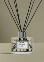 A reed diffuser bottle with black reeds, a clear glass body, and a silver cap, featuring a white label that reads 'ROSE DE MAI' and 'Rf Paris'. The background is a solid warm beige.