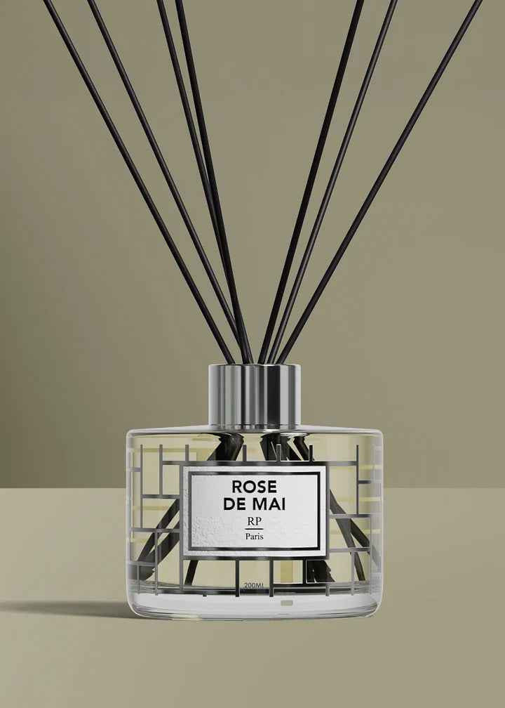 A reed diffuser bottle with black reeds, a clear glass body, and a silver cap, featuring a white label that reads 'ROSE DE MAI' and 'Rf Paris'. The background is a solid warm beige.