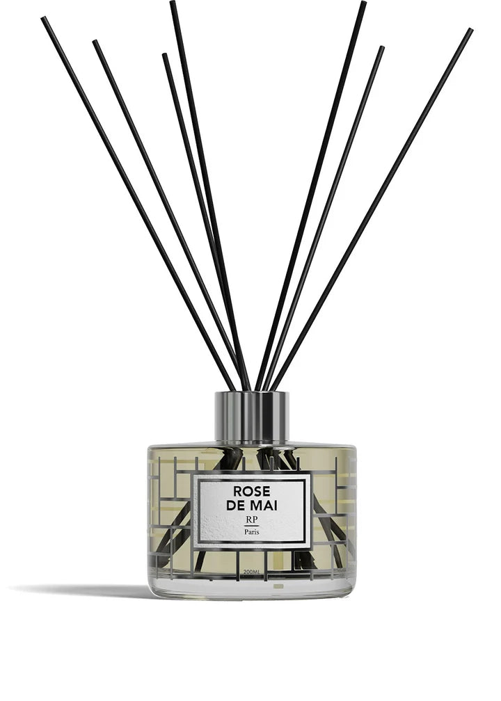 A reed diffuser bottle with a clear glass body, pale yellow liquid, a white label reading 'ROSE DE MAI BP Paris,' and multiple black reed sticks extending upward against a white background.