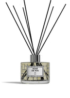 A reed diffuser bottle with a clear glass body, pale yellow liquid, a white label reading 'ROSE DE MAI BP Paris,' and multiple black reed sticks extending upward against a white background.