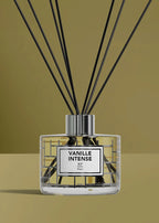 A reed diffuser bottle with black reeds, featuring a clear glass container with a white label that reads 'VANILLE INTENSE' and 'R.F.