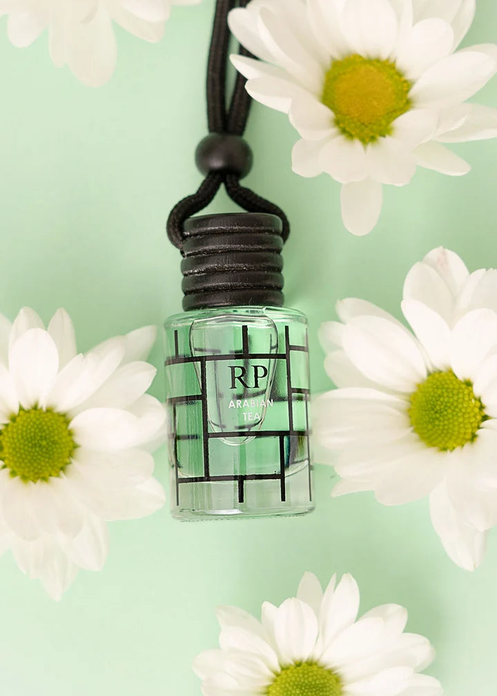 A small clear glass bottle with a black grid-patterned label and a black cord loop, centered on a pale mint green background and surrounded by white daisies.