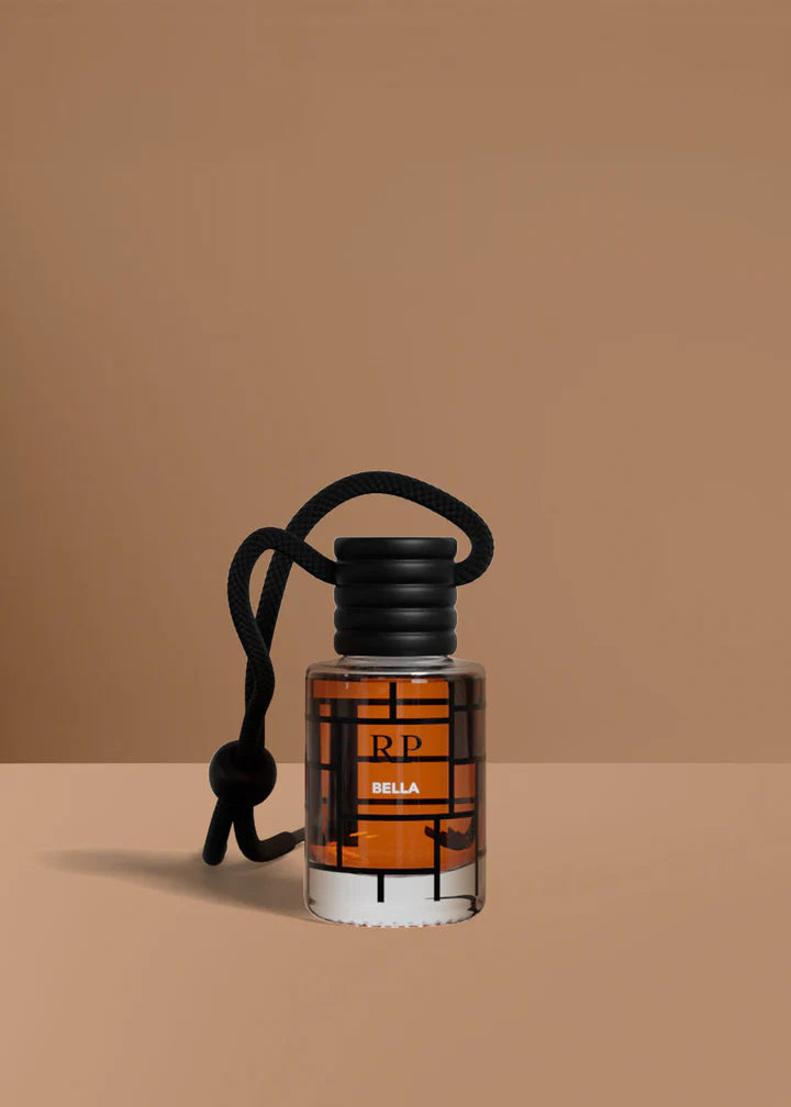 A small clear glass bottle with amber liquid and a black cord loop, labeled RP BELLA, set against a warm tan background.