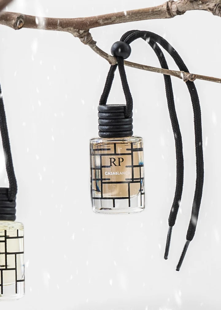 A small clear glass perfume bottle with a black cord hanger and black geometric pattern, hanging from a tree branch against a snowy white background. The bottle label reads RP and CASABLANA.