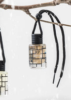 A small clear glass perfume bottle with a black cord hanger and black geometric pattern, hanging from a tree branch against a snowy white background. The bottle label reads RP and CASABLANA.
