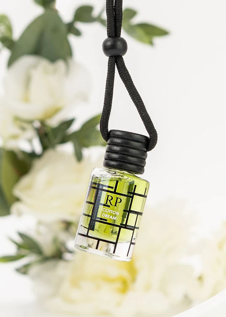 A small clear glass bottle filled with yellow-green liquid, hanging from a black cord. The bottle has a black grid pattern and a black screw cap, with the text 'RP COTTON DREAM' visible on the front.