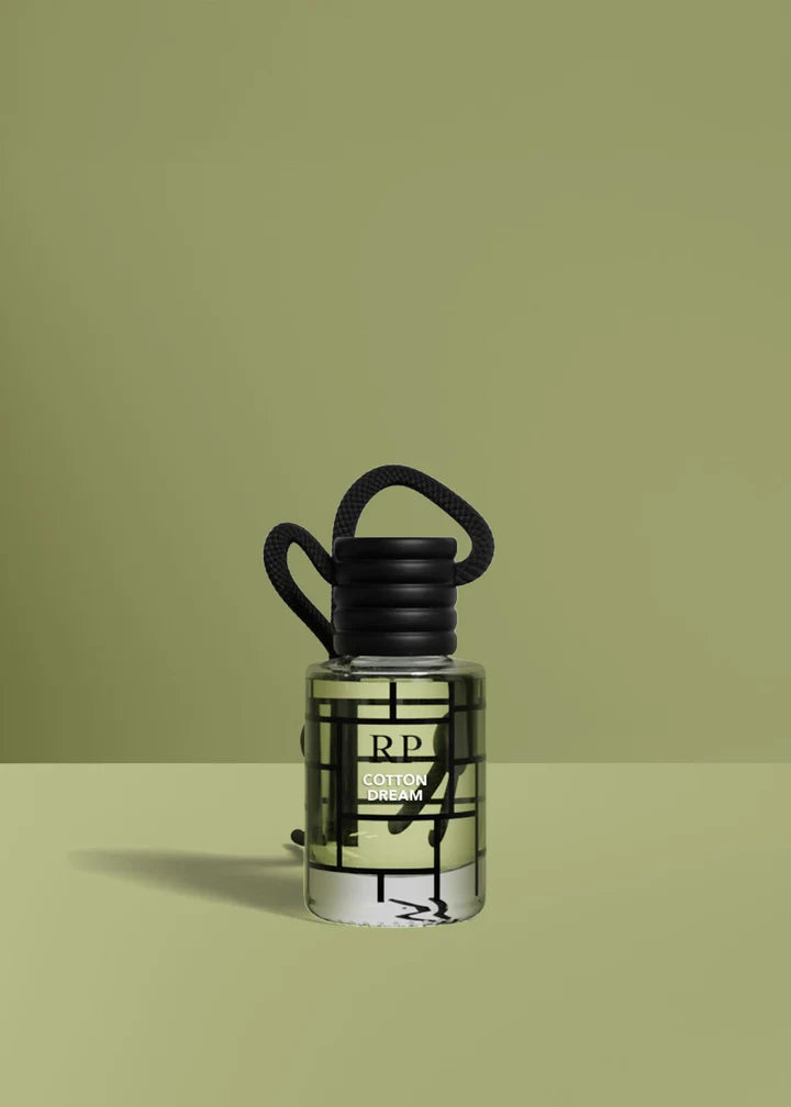 A small clear glass bottle with a black cap and black hanging loop, labeled RP cotton dream, centered on a muted olive-green studio background.