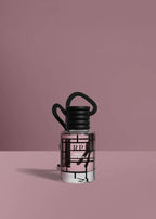 A small clear glass bottle with a black cap and black hanging loop, filled with pale pink liquid, set against a dusty pink background. The bottle has a black geometric grid pattern and a visible RP logo.FraiseDesBois
