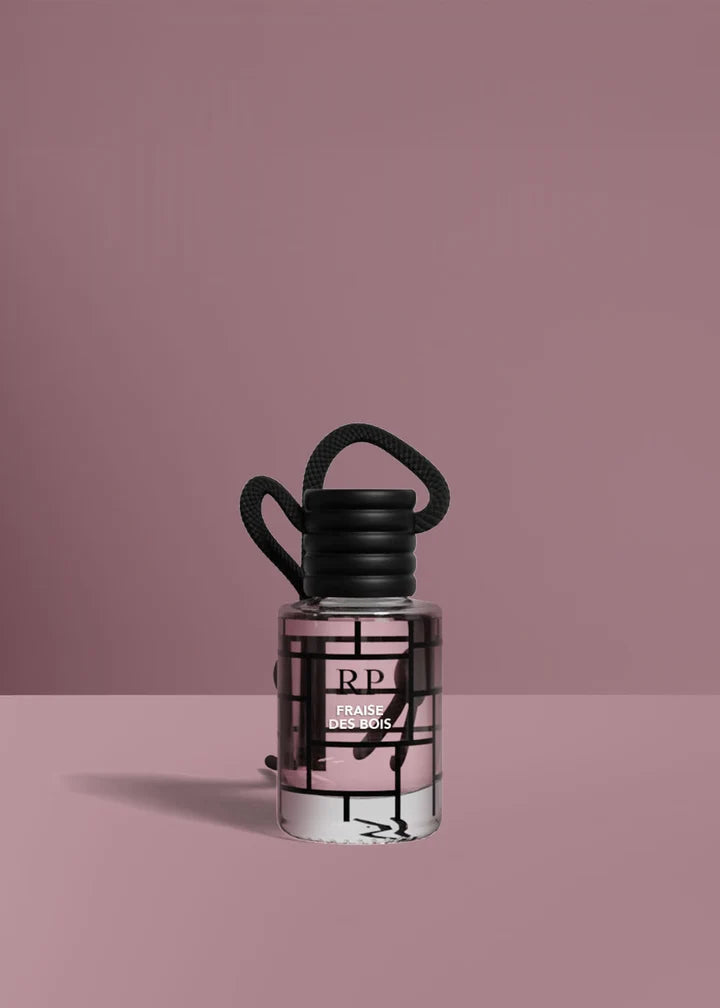 A small clear glass bottle with a black cap and black hanging loop, filled with pale pink liquid, set against a dusty pink background. The bottle has a black geometric grid pattern and a visible RP logo.FraiseDesBois