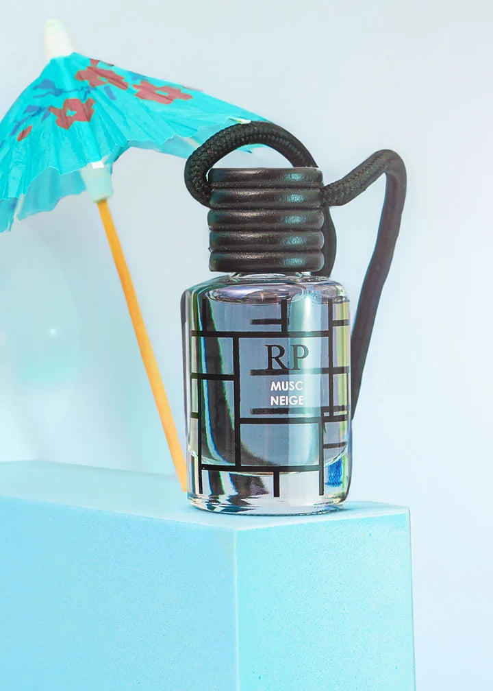 A clear glass bottle of RP Music Neige fragrance with a black coiled cap and dark handle, styled on a light blue pedestal with a turquoise cocktail umbrella prop against a pale blue background.