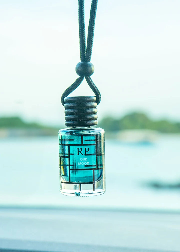 A small clear glass bottle with turquoise blue liquid and a black geometric pattern label, hanging from a black cord. The label reads 'RP' and 'oud MOOD'.