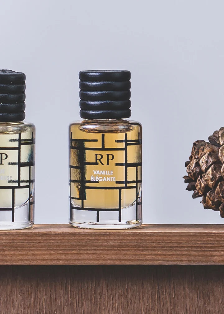 A small clear glass perfume bottle with a black ribbed cap and a black geometric label reading RP VANILLE ÉLÉGANT, shown on a wooden shelf next to another bottle and a pinecone against a light gray background.