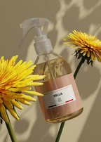 A clear plastic spray bottle with a white trigger sprayer and a blush pink label reading 'BELLA 100% Pure' and 'Belle France', styled with two yellow flowers against a beige background with soft leaf shadows.