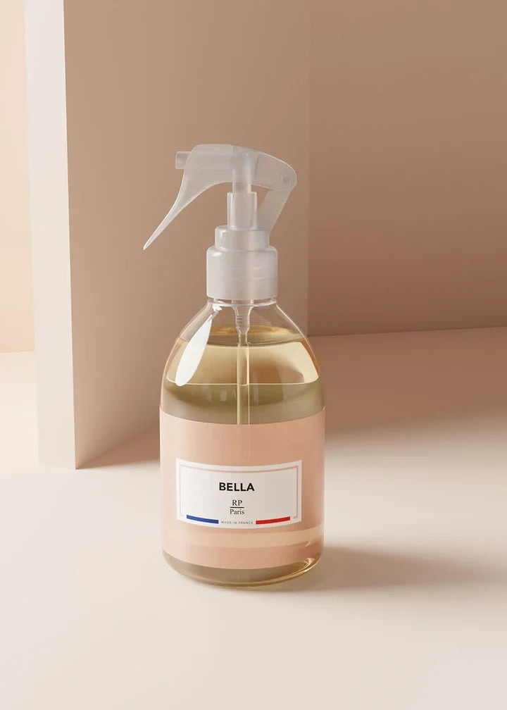 A clear trigger spray bottle filled with pale yellow liquid, featuring a soft blush pink label and a white spray nozzle. The bottle is labeled 'BELLA' and is set against a minimalist, warm beige background.