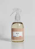 A clear spray bottle filled with pale yellow liquid, featuring a beige label with the brand name BELLA and a white trigger sprayer, set against a plain white background.