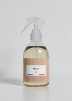 A clear spray bottle filled with pale yellow liquid, featuring a beige label with the brand name BELLA and a white trigger sprayer, set against a plain white background.