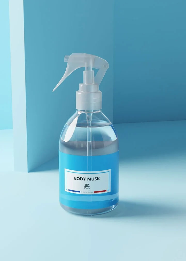 A clear plastic spray bottle filled with a light blue liquid, featuring a white trigger sprayer and a white label that reads 'BODY MUSK' with a small French flag detail, set against a minimalist blue background.