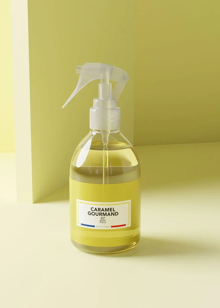 A clear plastic spray bottle filled with pale yellow liquid, featuring a white trigger sprayer and a white label that reads 'CARAMEL COUTMAND' with small blue and red accents. The bottle is set against a minimalist, solid pale yellow background.
