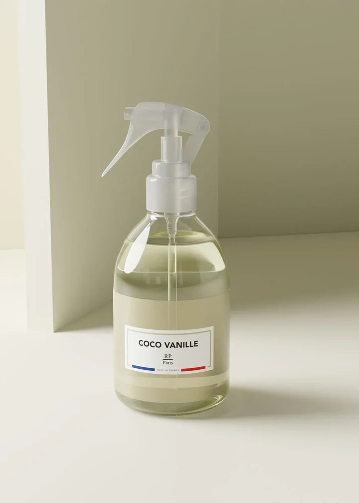 A clear glass spray bottle filled with a pale yellow liquid, featuring a white trigger sprayer and a white label that reads 'COCO VANILLE' with a small blue and red accent line, set against a minimalist beige background.