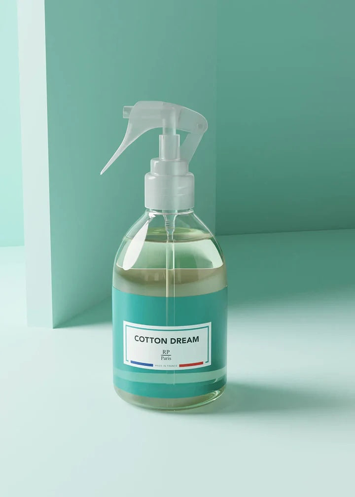 A clear glass spray bottle filled with a pale green liquid, featuring a white trigger sprayer and a teal label that reads 'COTTON DREAM' with a small French flag detail. The bottle is set against a minimalist, solid teal background.