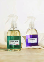 Two clear spray bottles of room spray with white triggers, one labeled 'COTTON DREAM' with a green label and the other 'MUSC BLANC' with a purple label, set on a light wooden surface with soft white towels in the background.