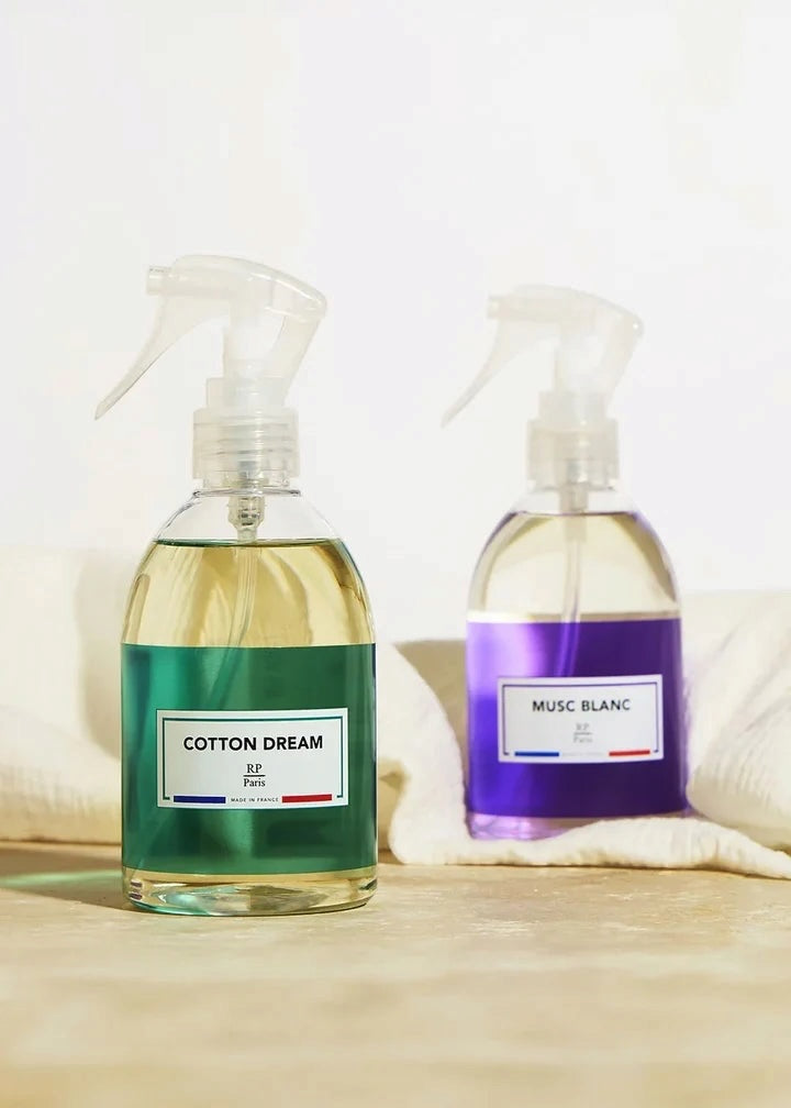 Two clear spray bottles of room spray with white triggers, one labeled 'COTTON DREAM' with a green label and the other 'MUSC BLANC' with a purple label, set on a light wooden surface with soft white towels in the background.