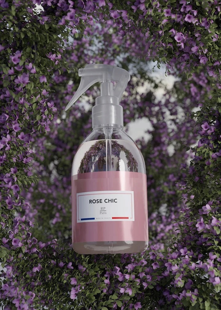 A clear plastic spray bottle with a pink label reading 'ROSE CHIC' and 'SP A/2' on the front, set against a background of dense purple flowers and green foliage.