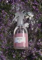 A clear plastic spray bottle with a pink label reading 'ROSE CHIC' and 'SP A/2' on the front, set against a background of dense purple flowers and green foliage.
