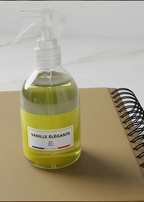 A clear plastic spray bottle filled with pale yellow liquid, featuring a white label that reads 'VANILLE ÉLÉGANTE' and a small French flag detail. The bottle is placed on a tan notebook next to a spiral-bound book on a light marble surface.