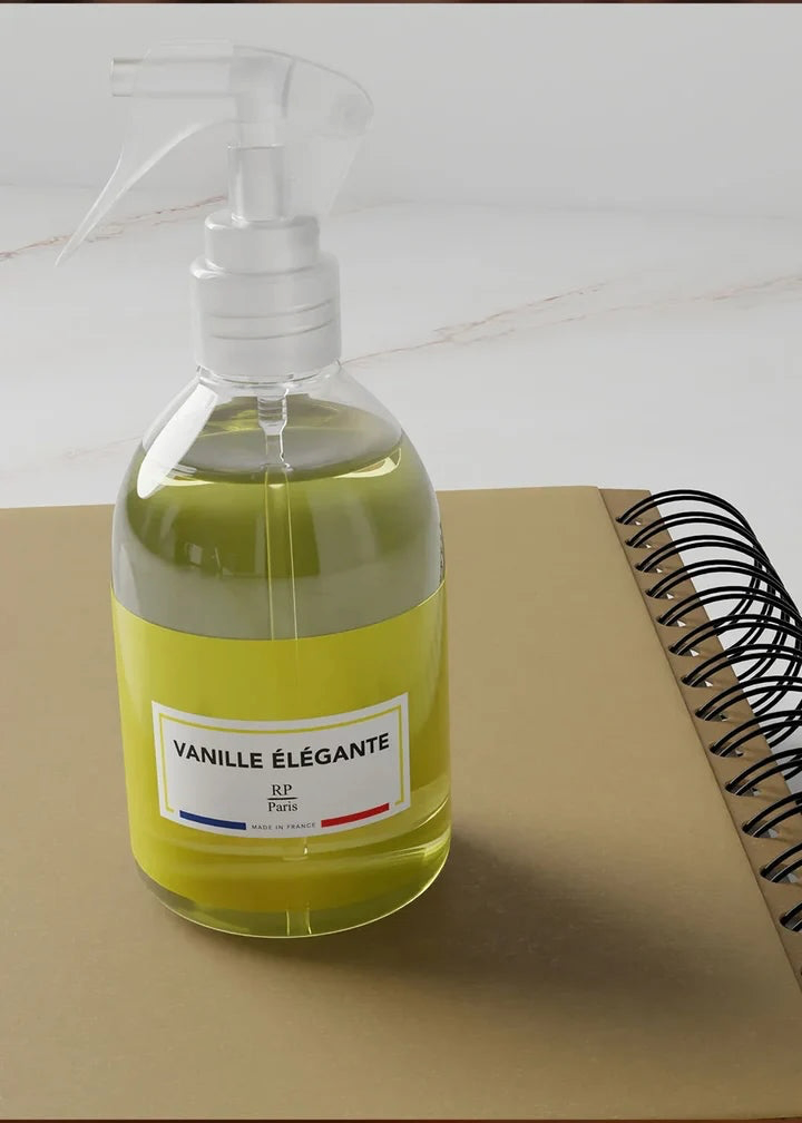 A clear plastic spray bottle filled with pale yellow liquid, featuring a white label that reads 'VANILLE ÉLÉGANTE' and a small French flag detail. The bottle is placed on a tan notebook next to a spiral-bound book on a light marble surface.