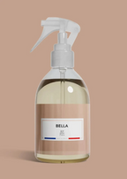 A clear plastic spray bottle filled with pale yellow liquid, featuring a white trigger sprayer and a beige label with the brand name BELLA and the text 'All Paris'. The bottle is centered against a solid, warm beige background.