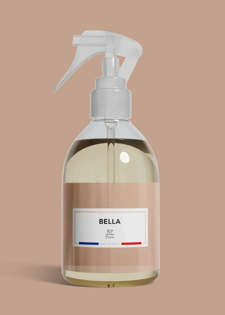 A clear plastic spray bottle filled with pale yellow liquid, featuring a white trigger sprayer and a beige label with the brand name BELLA and the text 'All Paris'. The bottle is centered against a solid, warm beige background.